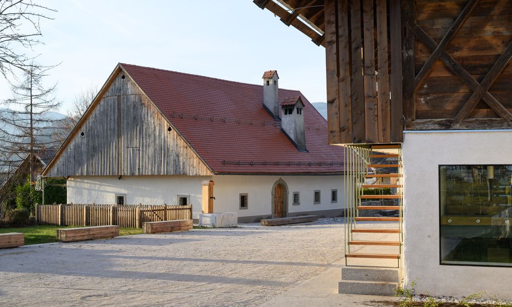 Pogled na prenovljeno Prešernovo hišo v Vrbi z lesenimi klopmi in pipo za vodo s kamnitim koritom.