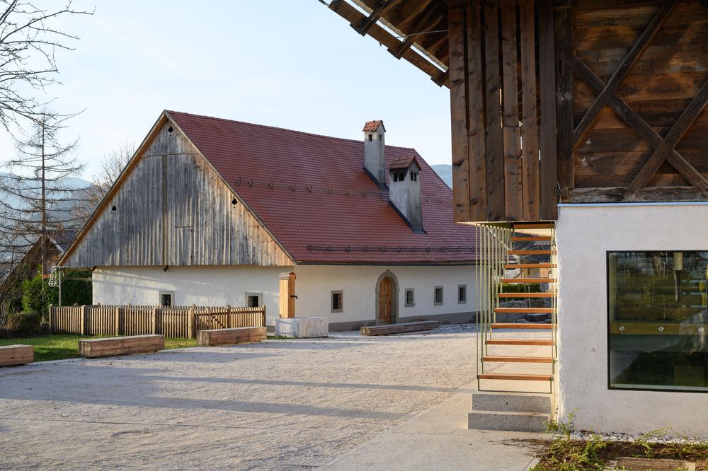 Pogled na prenovljeno Prešernovo hišo v Vrbi z lesenimi klopmi in pipo za vodo s kamnitim koritom.