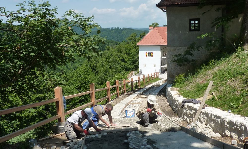 Trije delavci delajo na kamniti poti, obdani so z leseno ograjo na levi strani, ki vodi do gradu Podsreda.