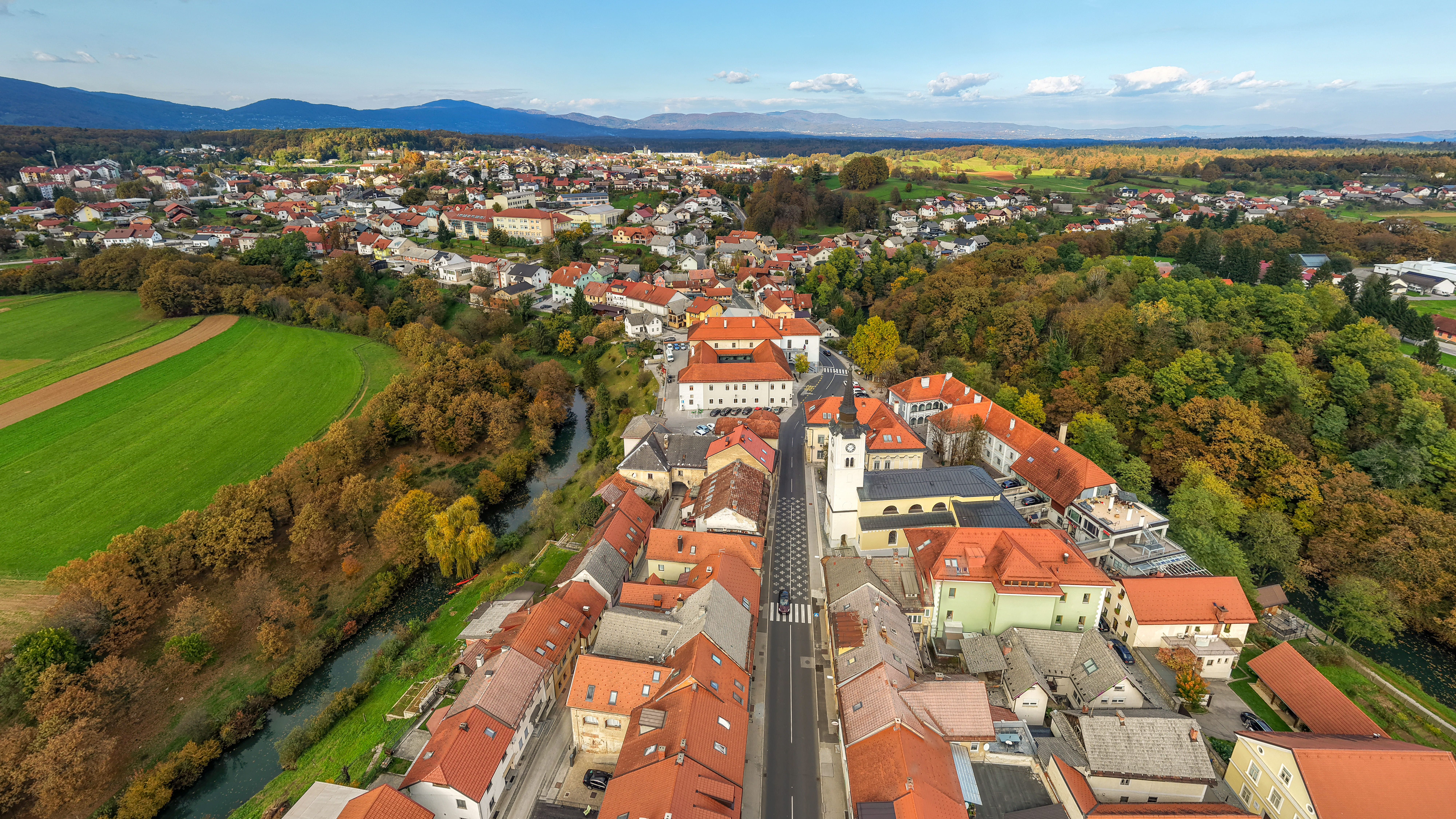 Grad Črnomelj iz zraka v daljavi, na straneh je obdan z zeleno-rjavim gozdom, spredaj in zadaj mesto zapolnijo oranžne strehe hi&scaron;