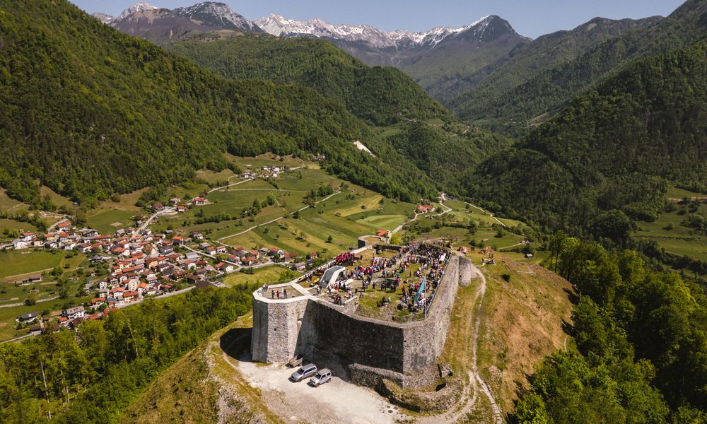Grad Kozlov rob je v ospredju, na njegovem travnatem vrhu stoji množica ljudi, v ozadju naselje Zatolmin ter pogled na Julijske Alpe.