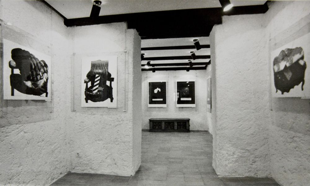 Črno-bela fotografija razstave v galeriji pred prenovo. Na belih stenah so umetniška dela.